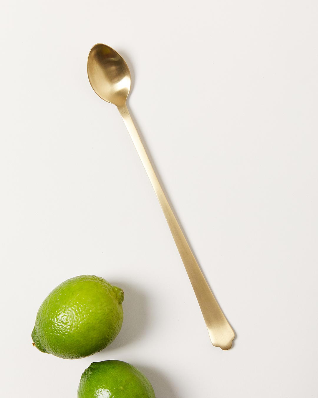 Long brushed-gold bar spoon displayed beside two whole limes, showing its small oval bowl and slender handle.