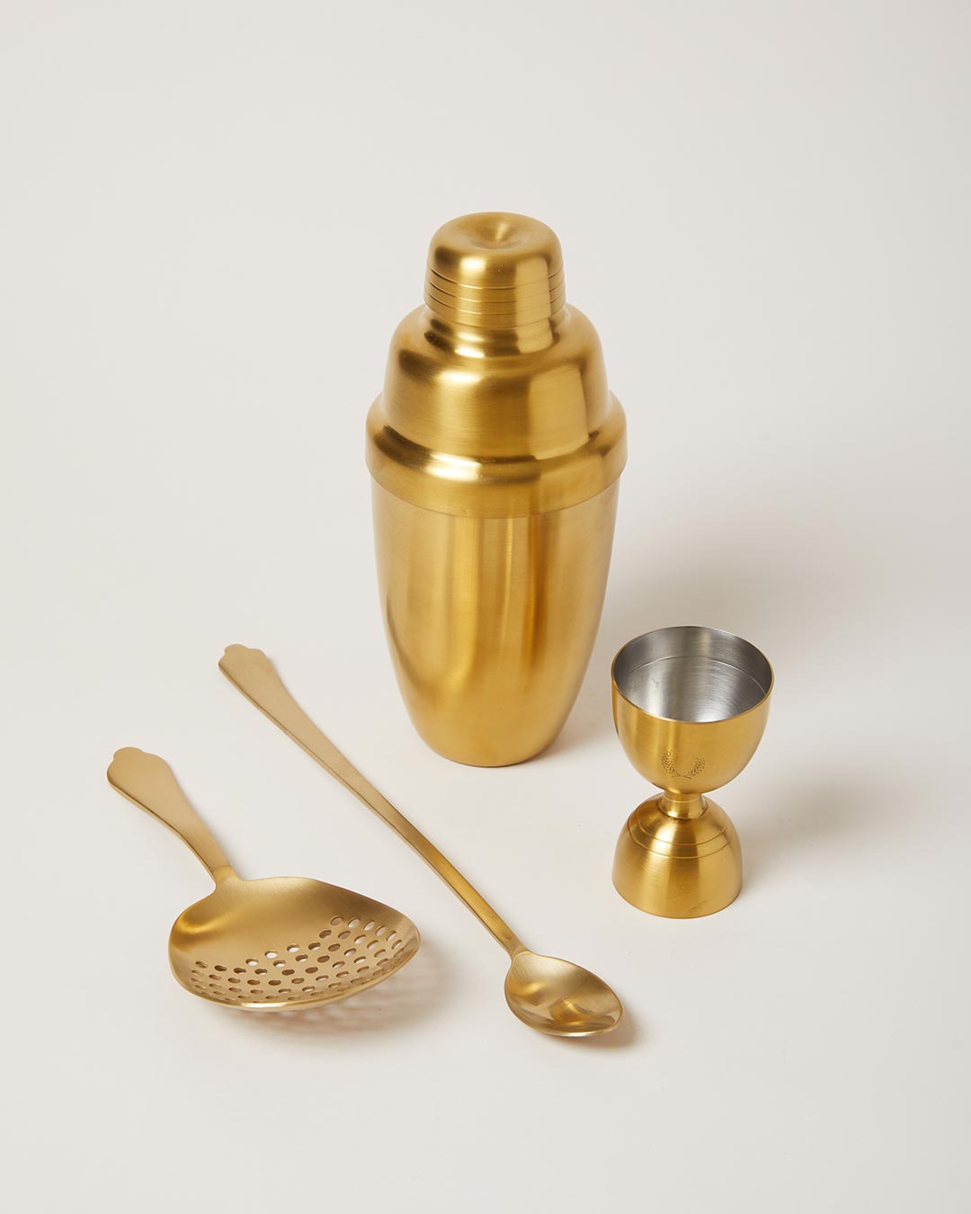 Four-piece brushed-gold barware set with a lidded cocktail shaker, double-sided jigger, perforated cocktail strainer, and long-handled bar spoon.