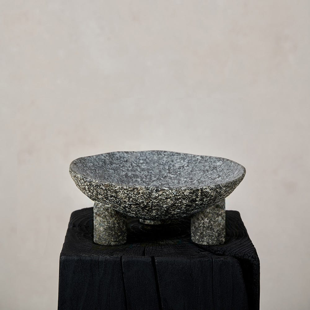 A carved stone footed bowl displayed on a black stand against a beige background.