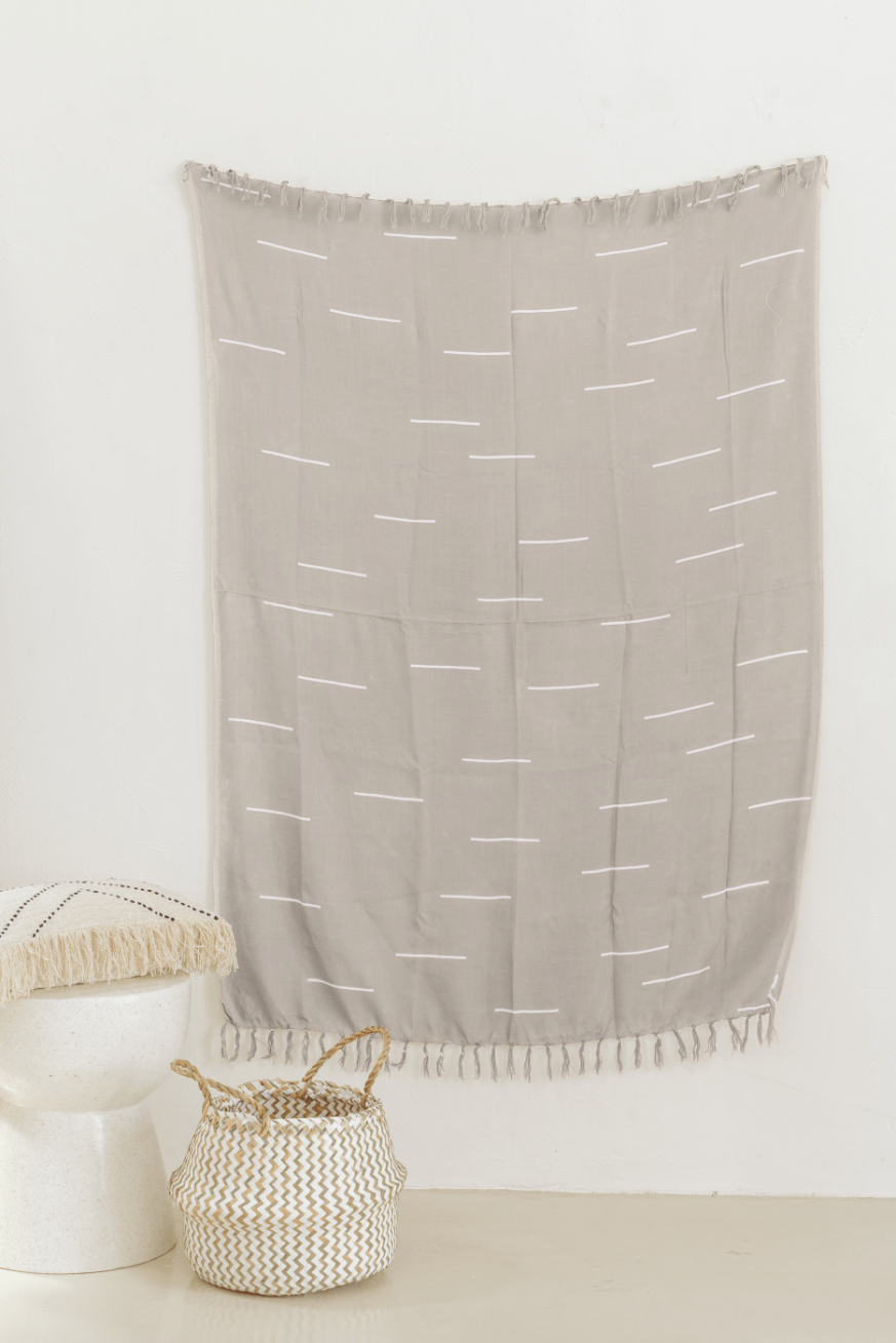 Village Thrive Lines Tapestry, taupe rayon with white line print, hanging on a wall with a wooden bench and basket below.