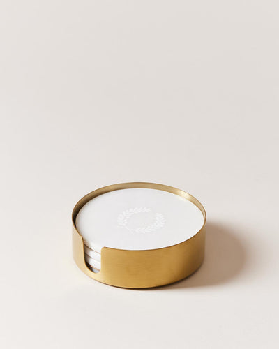 Round white marble coasters stacked inside a low cylindrical brass holder, with the top coaster engraved with a delicate white laurel wreath.