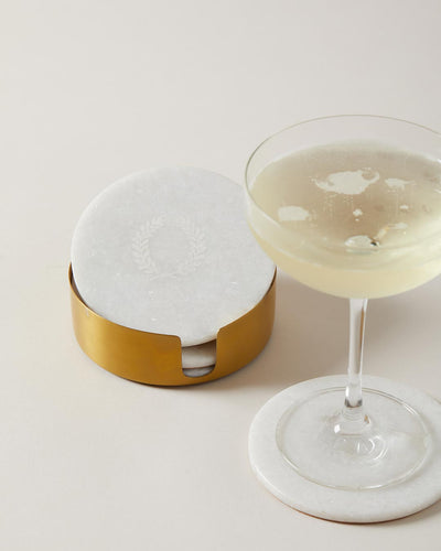 Coupe glass filled with a pale cocktail resting on a white marble coaster beside the brass coaster holder.