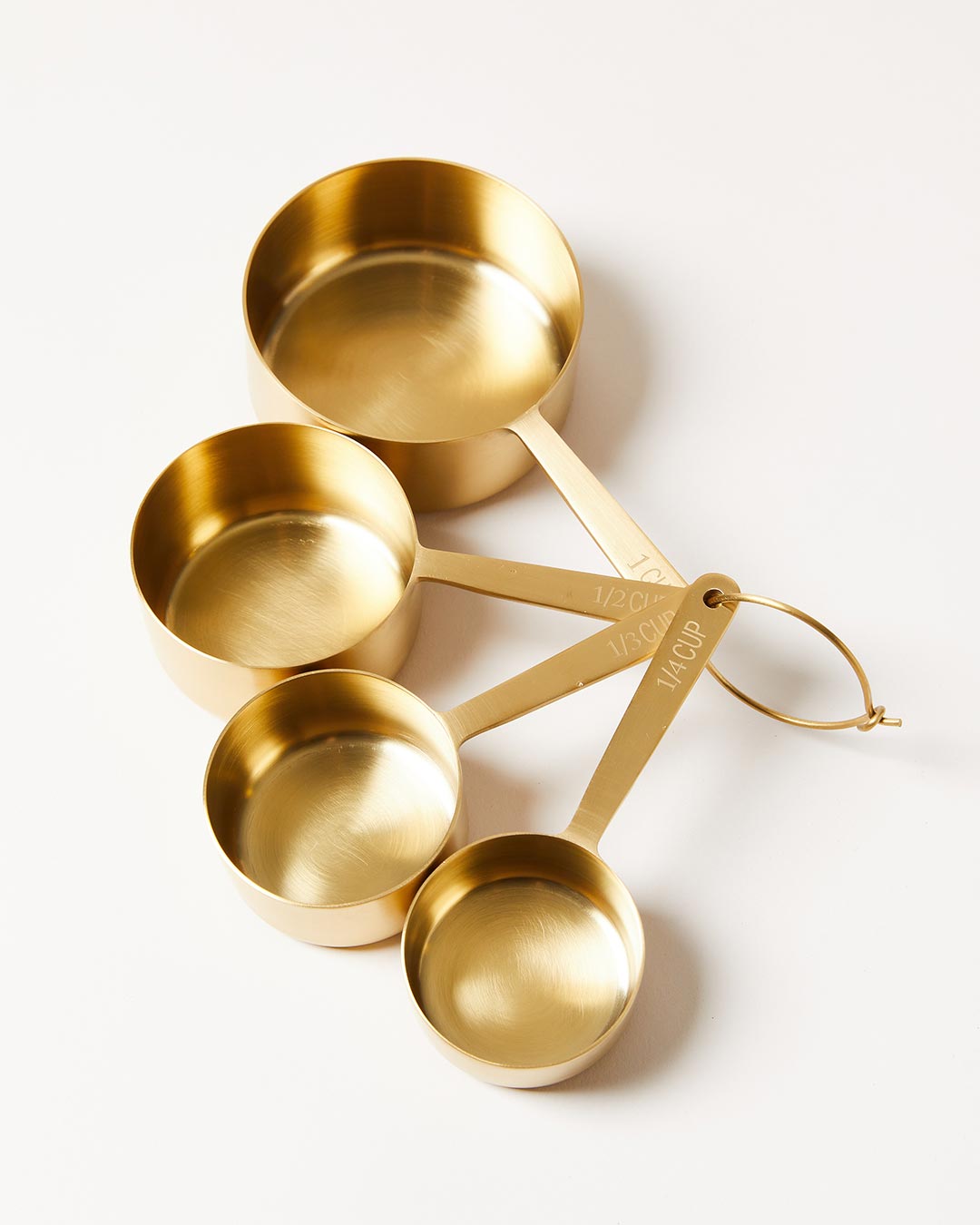 Four brushed brass measuring cups fanned outward from a small gold storage ring on a pale neutral surface.