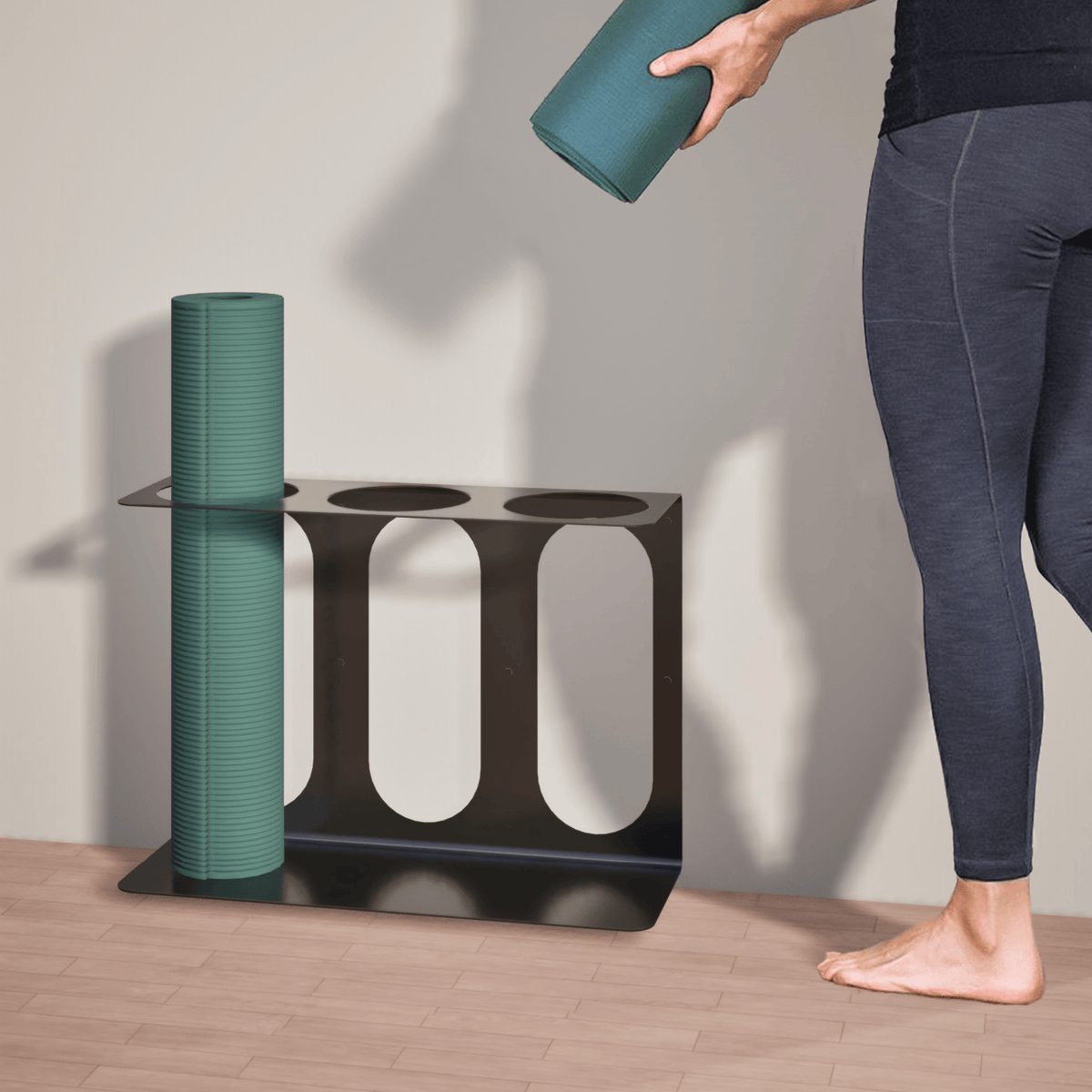 Black metal wall bracket with three vertical storage slots, shown holding one teal foam roller as a person lifts a second roller toward an open slot.