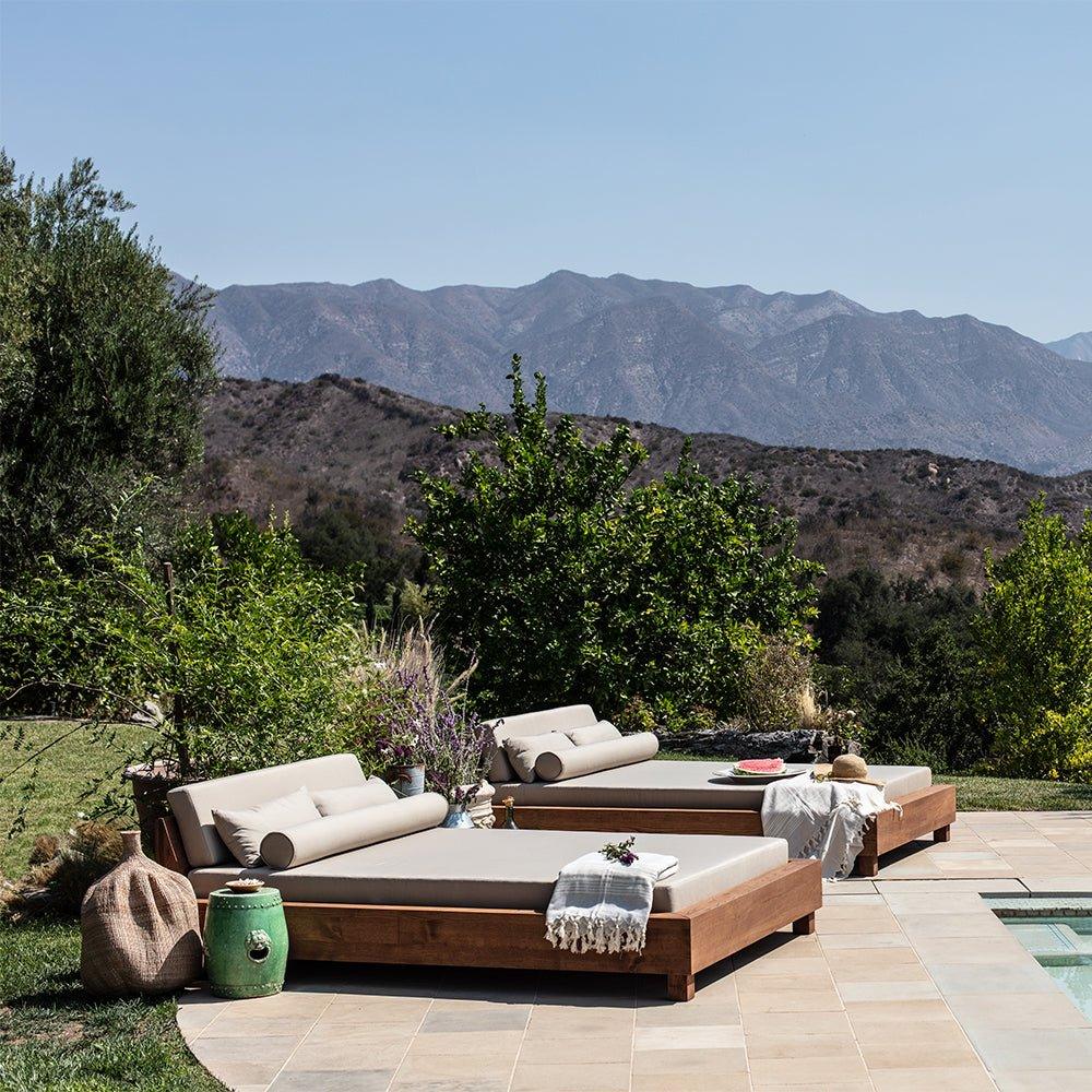 An Ojai Outdoor Daybed with cushions, placed on a tiled area next to a pool with a mountainous landscape in the background.