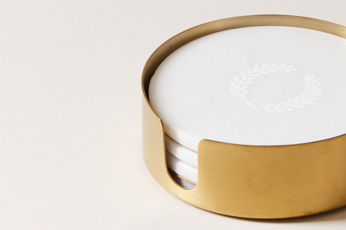 Close-up of the brass holder filled with white marble coasters, showing subtle gray veining, polished edges, and the engraved laurel wreath.
