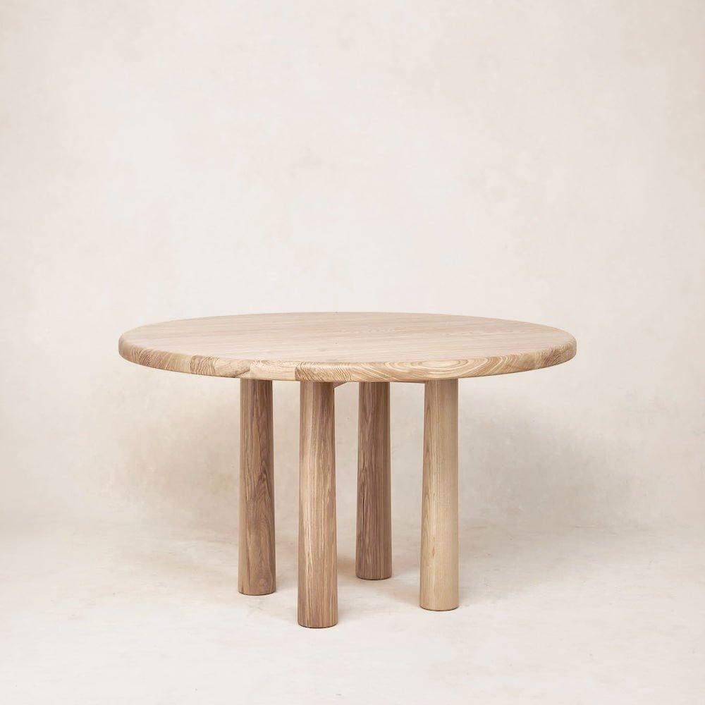 A round wooden dining table with a natural finish and four cylindrical legs, displayed against a neutral background.