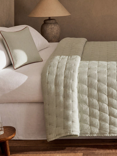 Pale sage green quilt draped across white bedding beside a matching accent pillow, with its pale reverse visible along the folded edge.