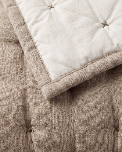 Close-up of the taupe quilt’s textured fabric, tufted stitches, and bound edge beneath a folded cream reverse.
