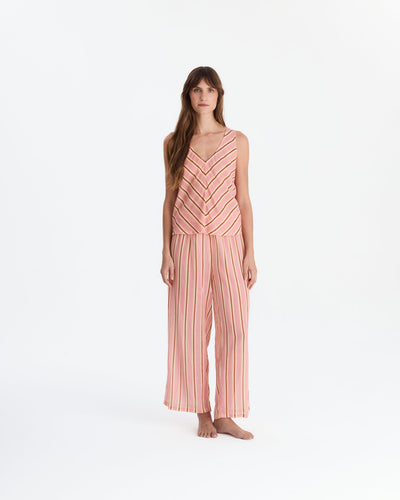 Barefoot model wearing coral pink pajamas with a sleeveless V-neck top featuring chevron stripes and matching vertically striped, wide-leg pants.