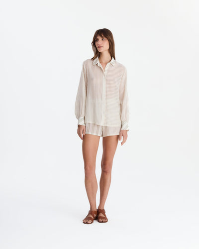 Model wearing cream and beige striped pajamas with a loose long-sleeve collared shirt, contrasting stripe directions, matching shorts, and brown sandals.