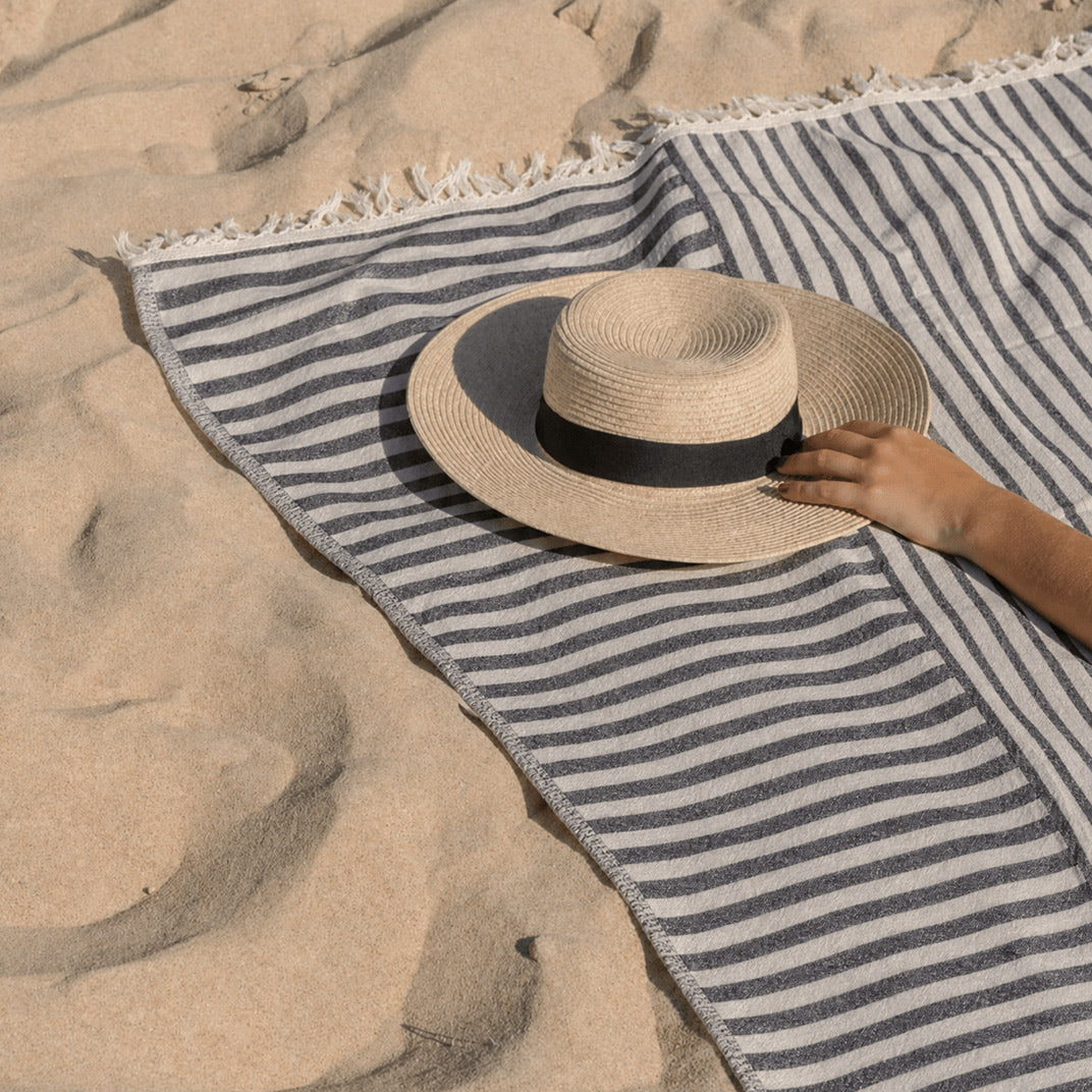 Striped Riva blanket spread across sand, with a straw hat resting on top and a person’s hand touching its brim.