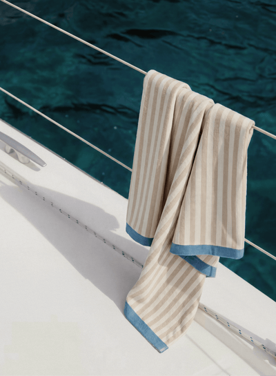 Beige and cream striped towel draped over a boat’s railing above deep blue water, with its blue-edged ends hanging over the white deck.