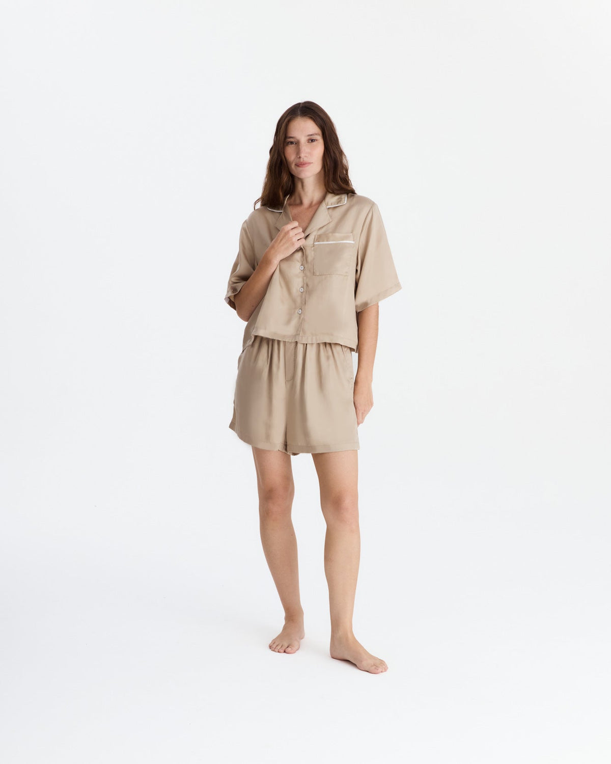 Full-length front view of a barefoot model wearing a beige pajama set with a loose short-sleeve button-front shirt, notched collar, white-trimmed chest pocket, and matching shorts.