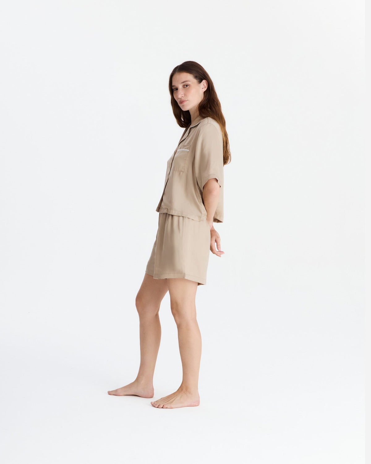 Side view of the beige pajama set on a barefoot model, showing the shirt’s loose drape, wide short sleeves, and matching upper-thigh-length shorts.