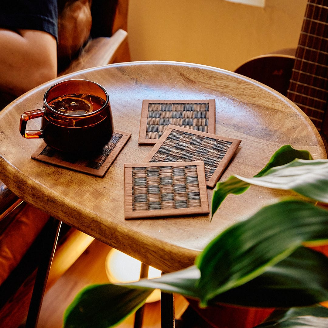 Wooden table with a mug of coffee and woven coasters, surrounded by greenery.
