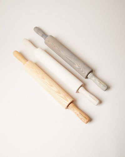 Three traditional ash rolling pins arranged diagonally in natural honey, whitewashed, and gray-washed finishes, each with a wide barrel and carved handles.