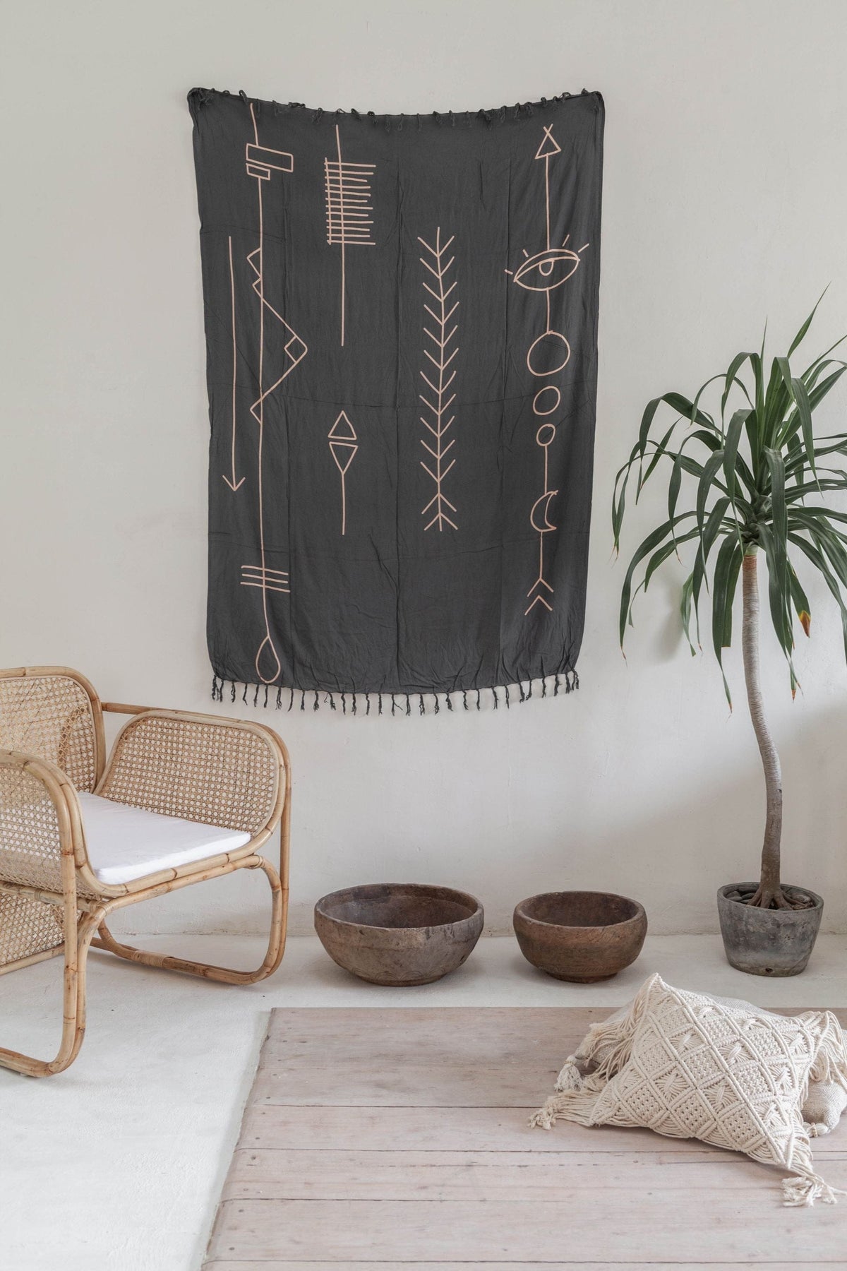 Village Thrive Abstract Tapestry, charcoal rayon, hanging above a rattan chair with a potted plant and floor cushion.