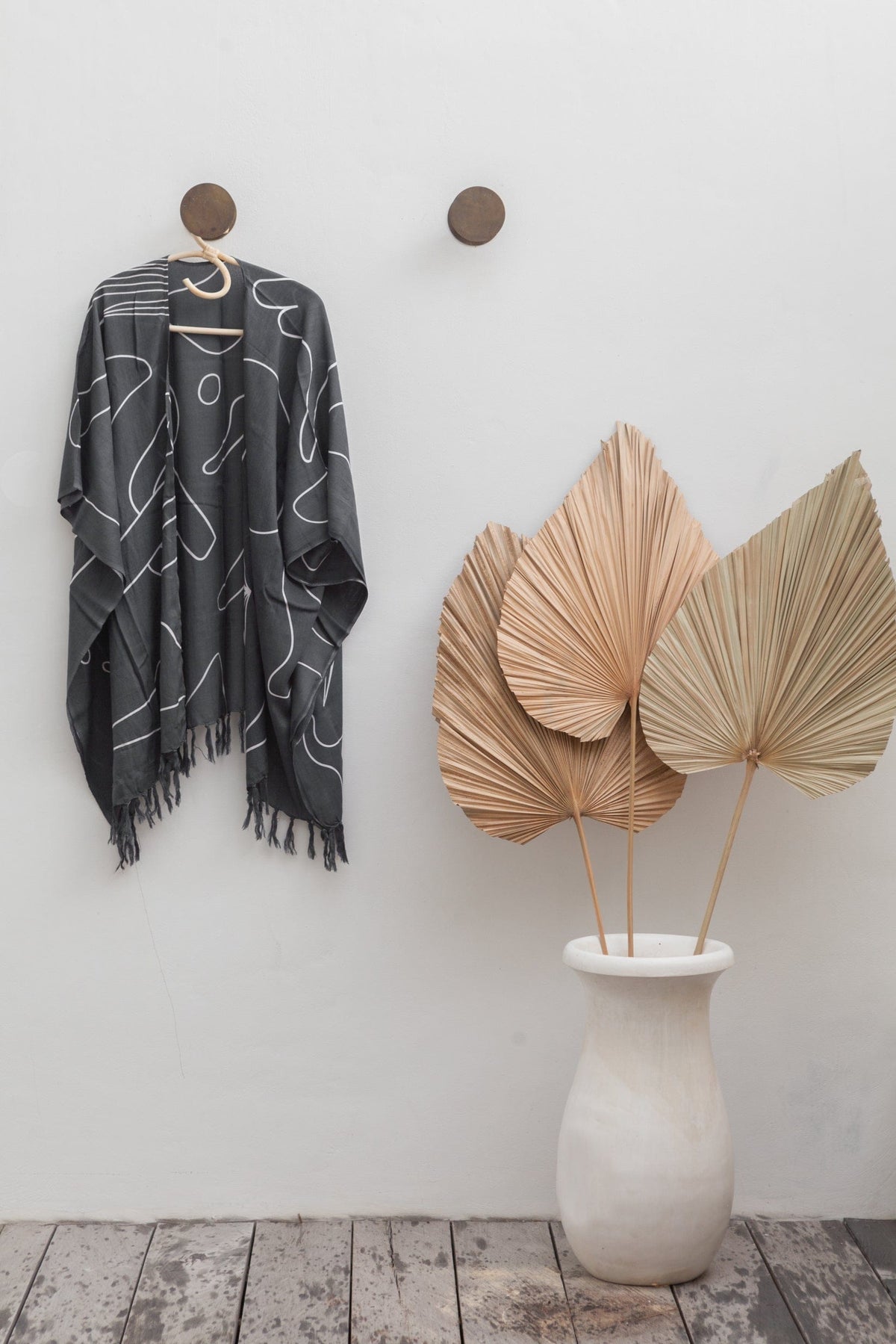 Village Thrive Desert Kimono, black rayon, hanging from a wall hook beside dried palm fronds in a ceramic vase.
