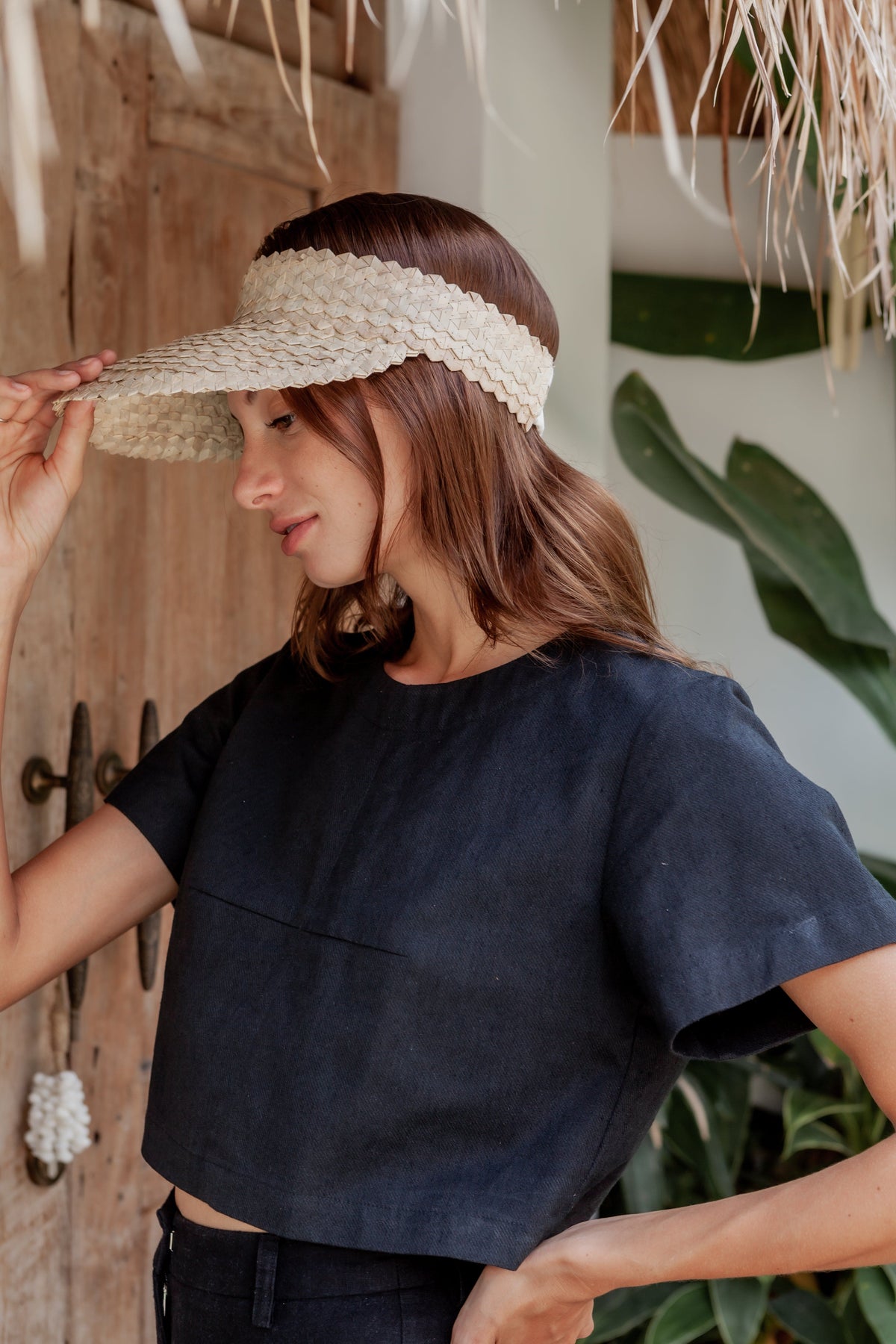 Village Thrive Rattan Sun Visor, blonde palm leaf, worn by a model adjusting the brim, standing by a wooden door.